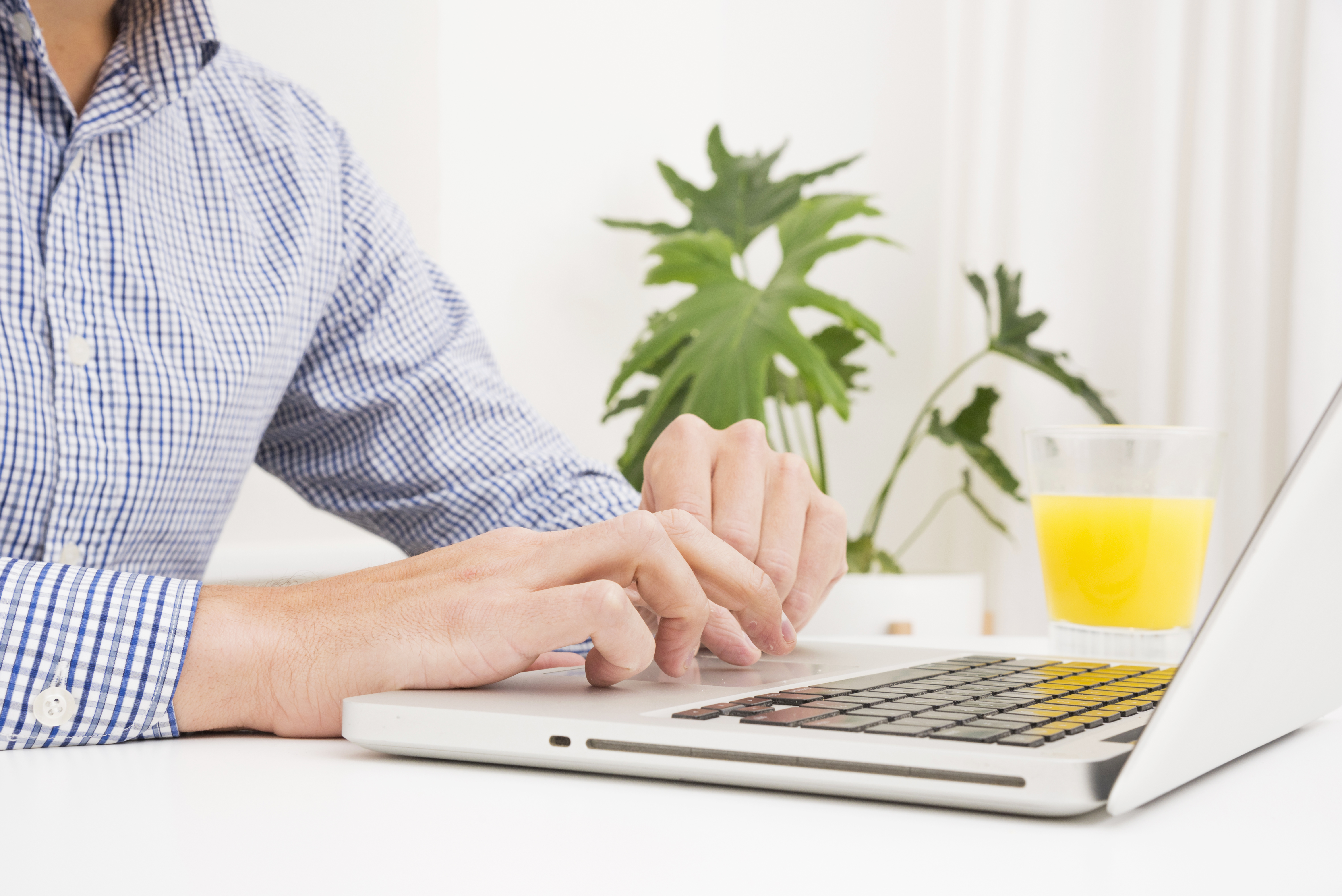 un homme en chemise bleue tape sur le clavier d'un ordinateur portable, une plante et un verre de jus d'orange à l'arrière plan.
